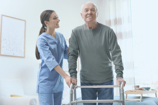Older man smiling and using frame after hip surgery with help of nurse at home